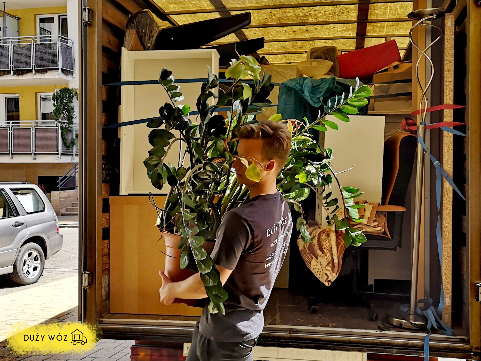 Mężczyzna w szarej koszulce z logo firmy przeprowadzkowej niesie dużą roślinę doniczkową z wnętrza furgonetki na tle załadowanych mebli i budynku mieszkalnego.
