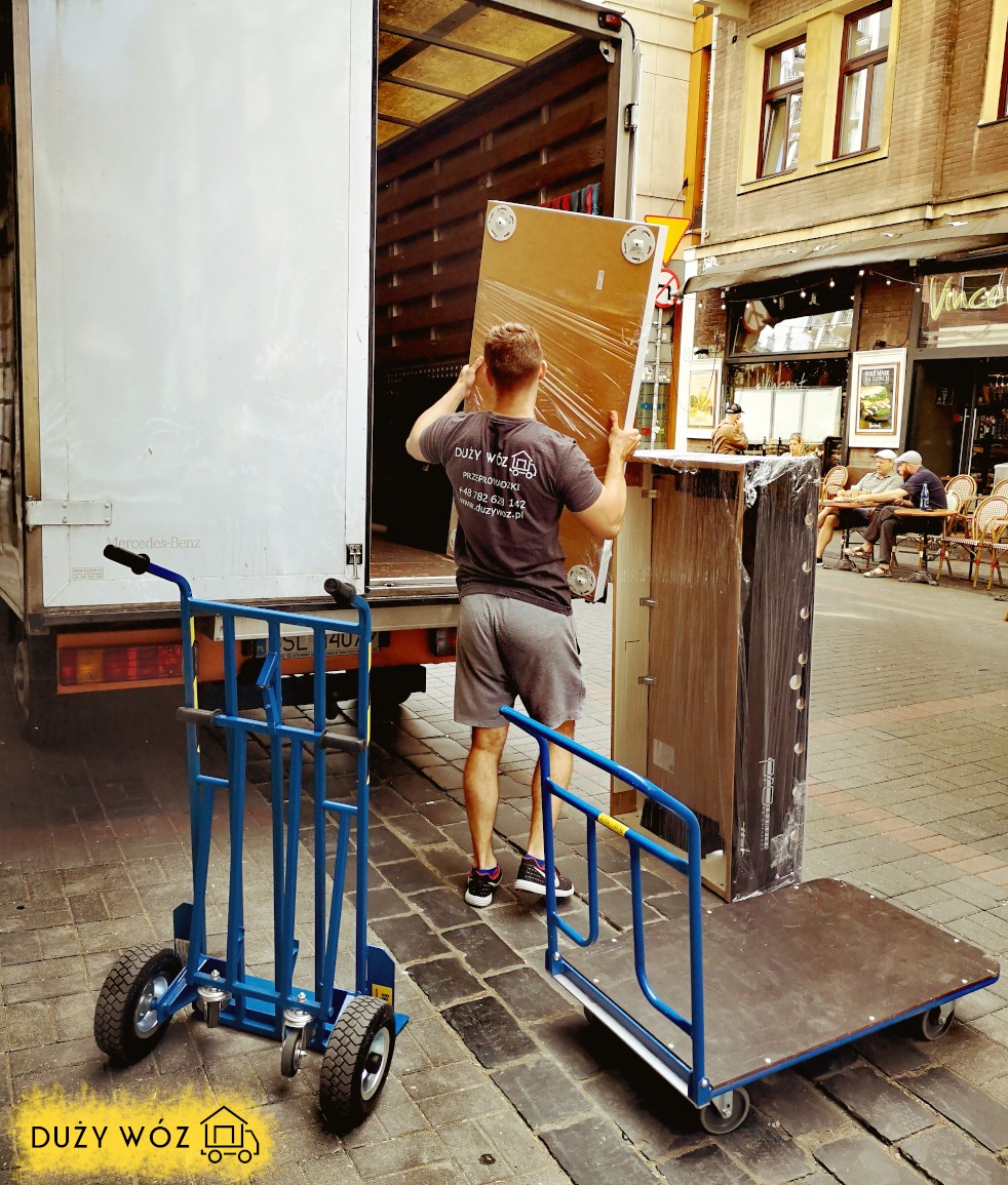Pracownik firmy przeprowadzkowej w szarej koszulce z logo wnosi zapakowaną płytę meblową do wnętrza białej ciężarówki, w tle widoczne wózki transportowe i ulica z kawiarnią.