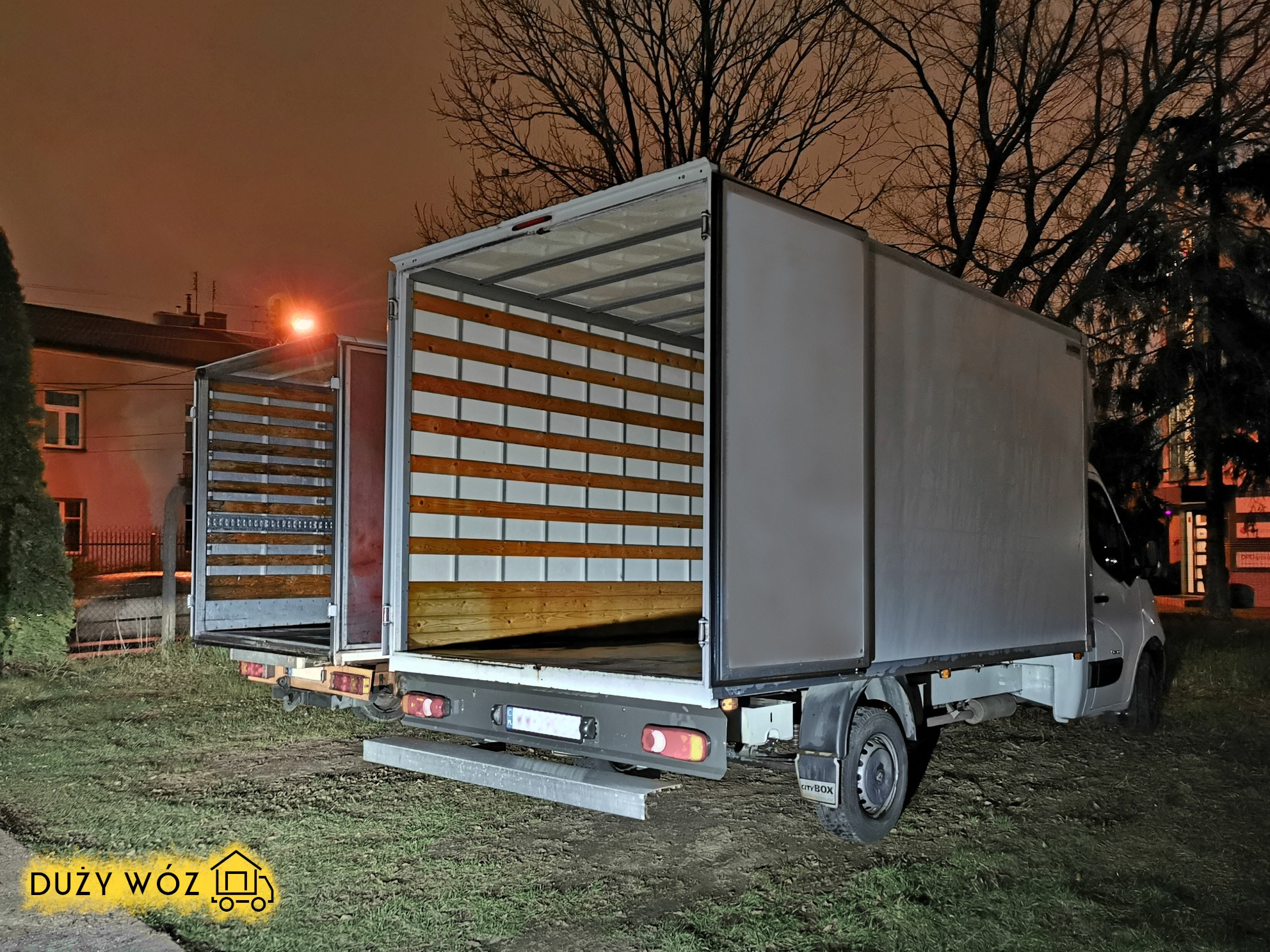 Otwarty, biały samochód dostawczy z drewnianą podłogą i ścianami bocznymi, stojący na trawie w nocy pod latarnią.