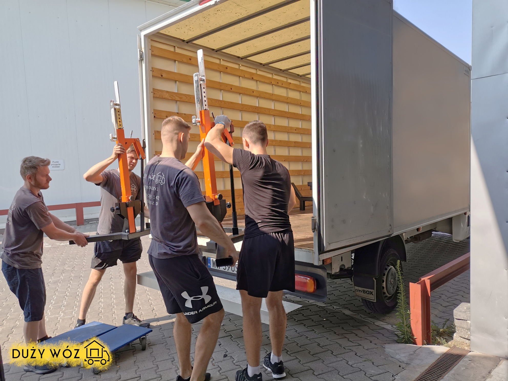 Czterech mężczyzn w trakcie załadunku mebli do furgonetki za pomocą specjalistycznych podnośników, widoczny wózek transportowy i logo firmy na dole zdjęcia.