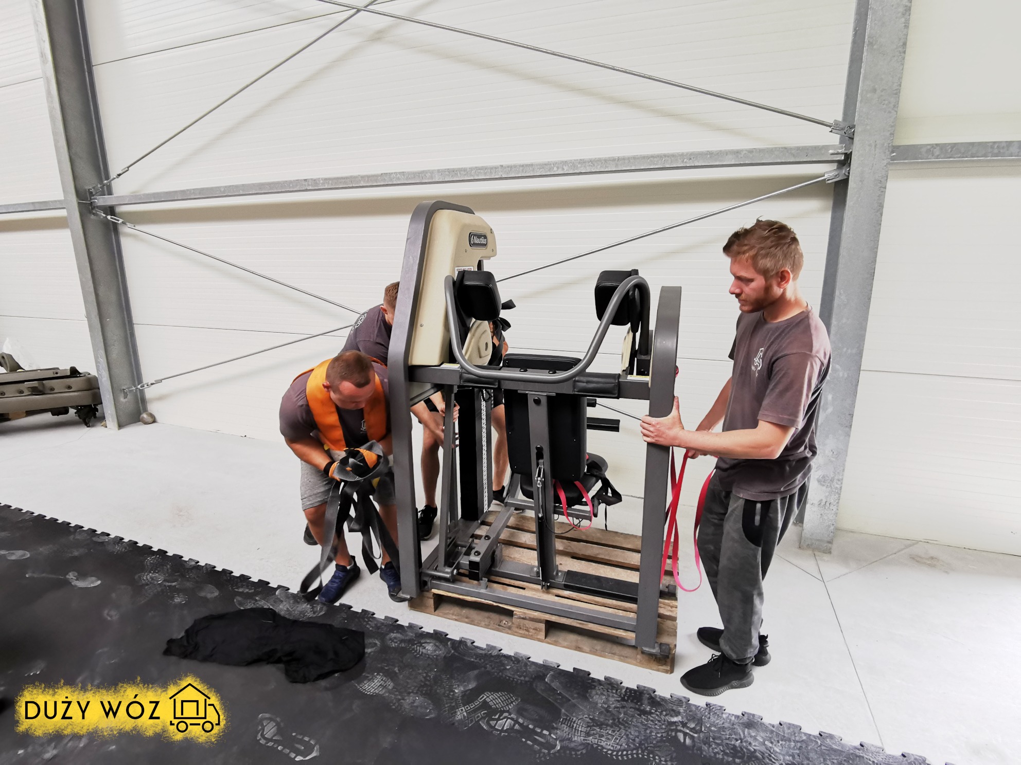 Trzech mężczyzn przenosi dużą maszynę do ćwiczeń, zapakowaną na palecie, wewnątrz magazynu z widocznym logo firmy transportowej na podłodze