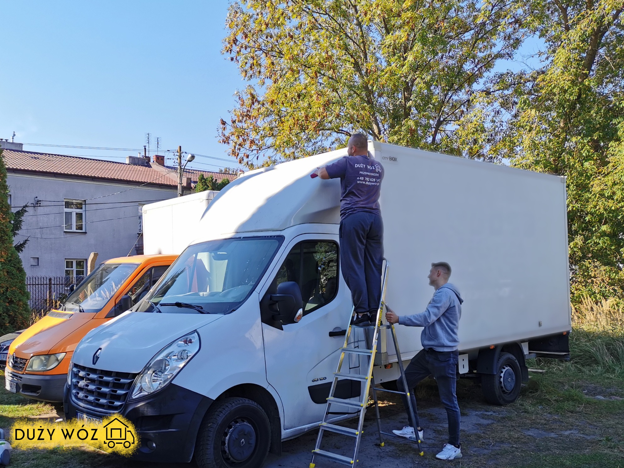 Dwaj mężczyźni oklejają białą furgonetkę z nadrukowanym logo firmy przeprowadzkowej, jeden stoi na drabinie, w tle pomarańczowy bus i budynek mieszkalny.