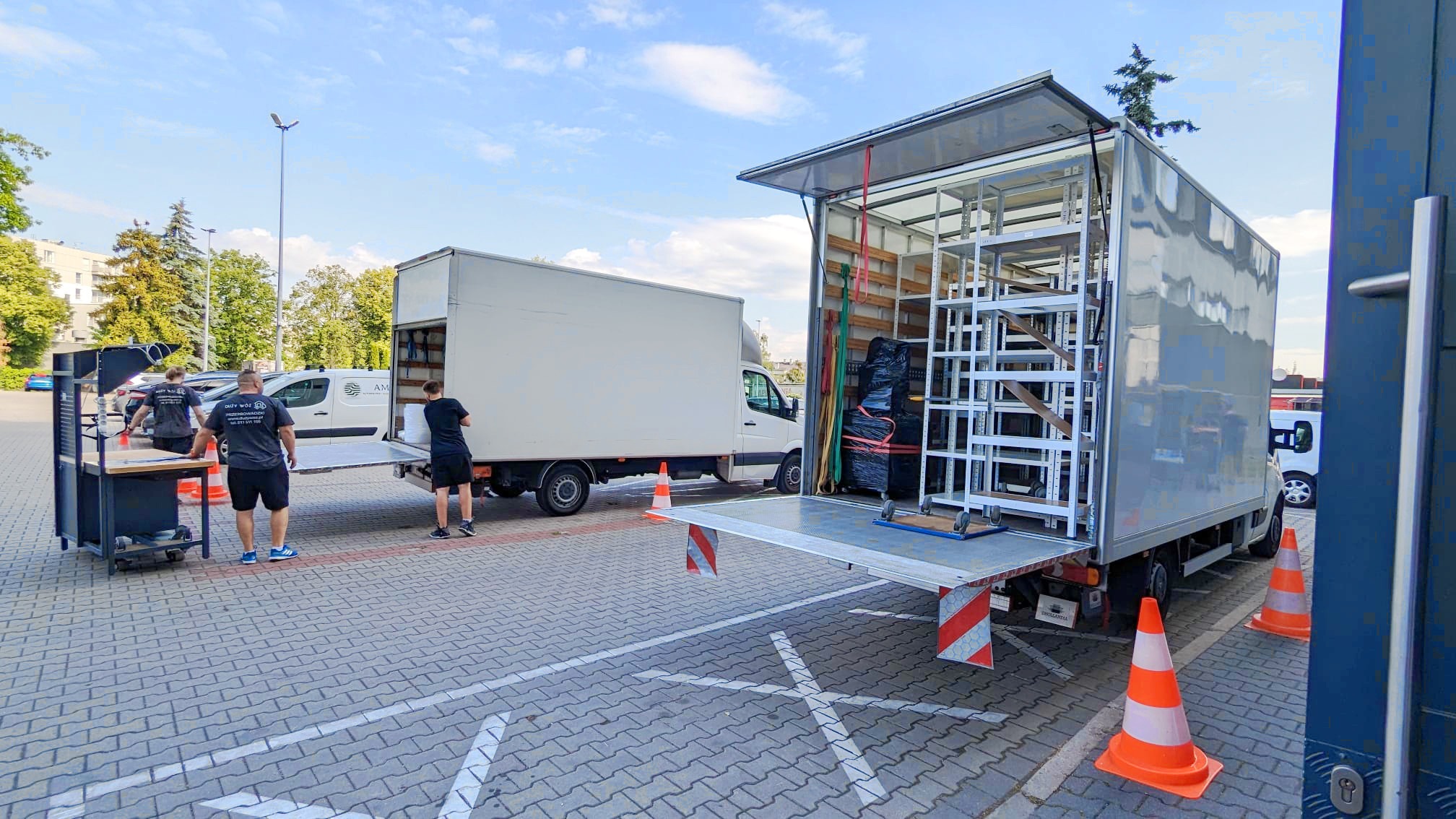 Dwa busy transportowe na parkingu z kostki brukowej. Jeden bus z otwartą rampą załadunkową i metalową konstrukcją wewnątrz. Pracownicy w czarnych koszulkach i spodenkach przygotowują się...