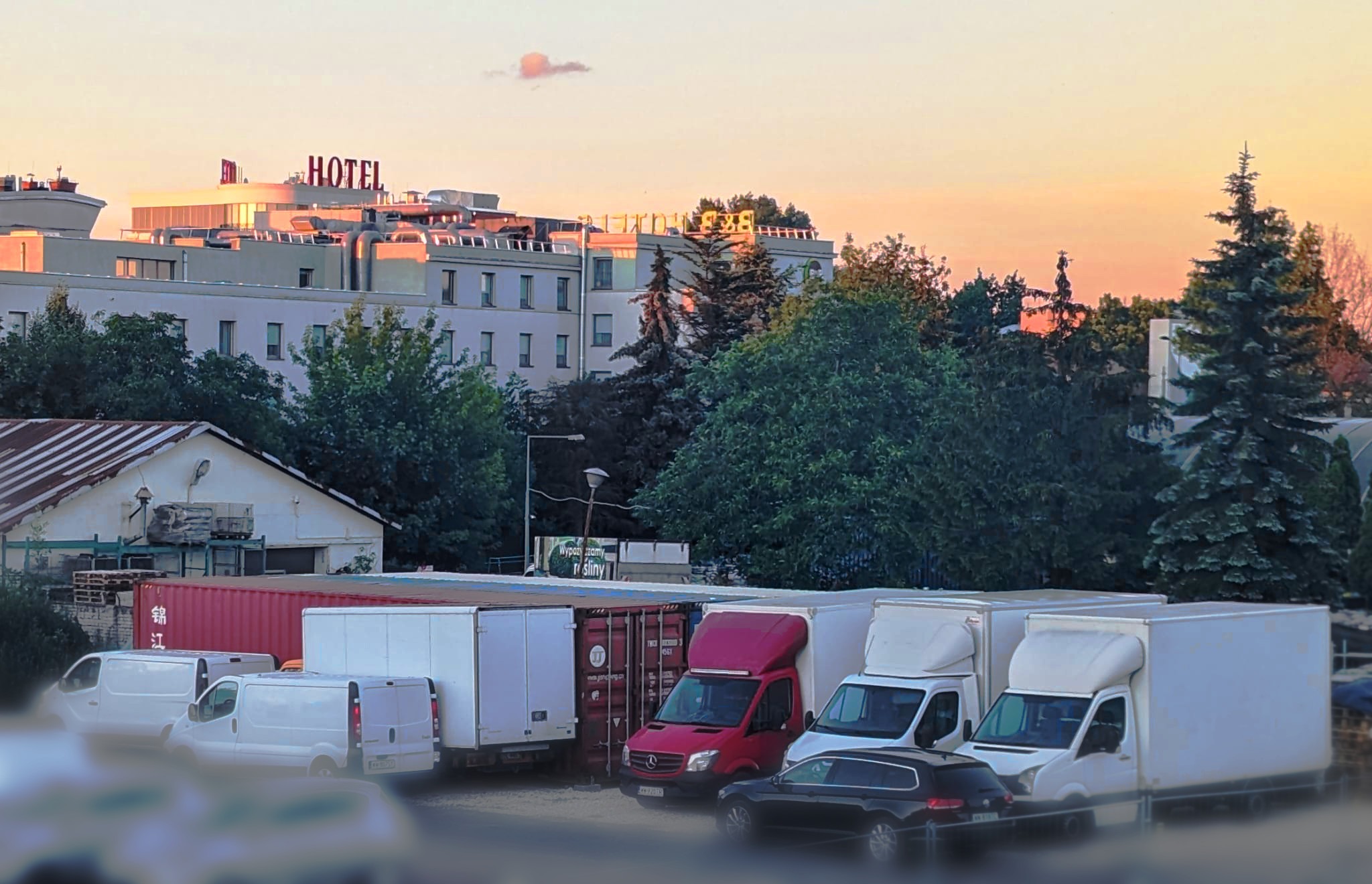 Flota transportowa: białe furgonetki i czerwony bus Mercedes, zaparkowane przed budynkiem z napisem HOTEL na dachu, w tle drzewa i zachodzące słońce.