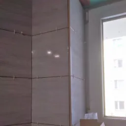 Świeżo położone, błyszczące, beżowo-szare płytki w rogu łazienki, z widocznymi krzyżykami dystansowymi i oknem w tle, w jasnym świetle.