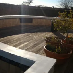 Półokrągły taras z drewnianą podłogą i niskim murem z jasnego kamienia, na którym widnieją napisy, z donicami i małymi drzewkami oraz krzesłami w tle, oświetlony słońcem.