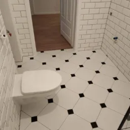 Nowoczesna łazienka z białymi, prostokątnymi płytkami na ścianach i podłodze w biało-czarną szachownicę; wisząca toaleta z deską; widok na otwarte drzwi do innego pomieszczenia z drewnianą podłogą.