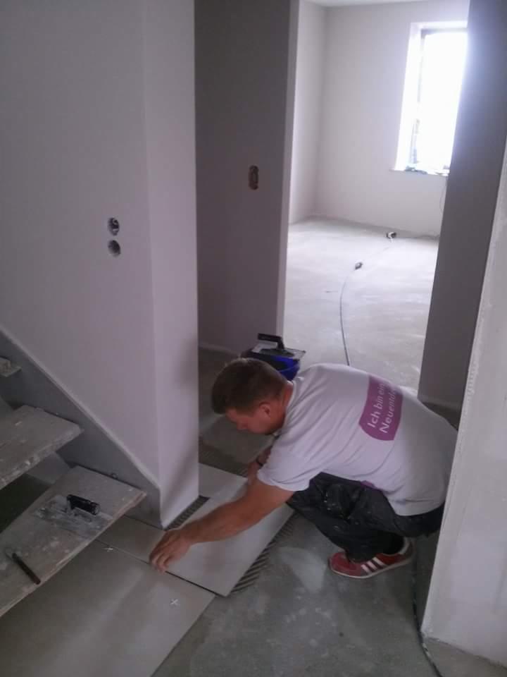 Mężczyzna w białej koszulce z krótkim rękawem, układający szare płytki pod schodami w trakcie remontu. Widoczne krzyżyki dystansowe i narzędzia glazurnicze.