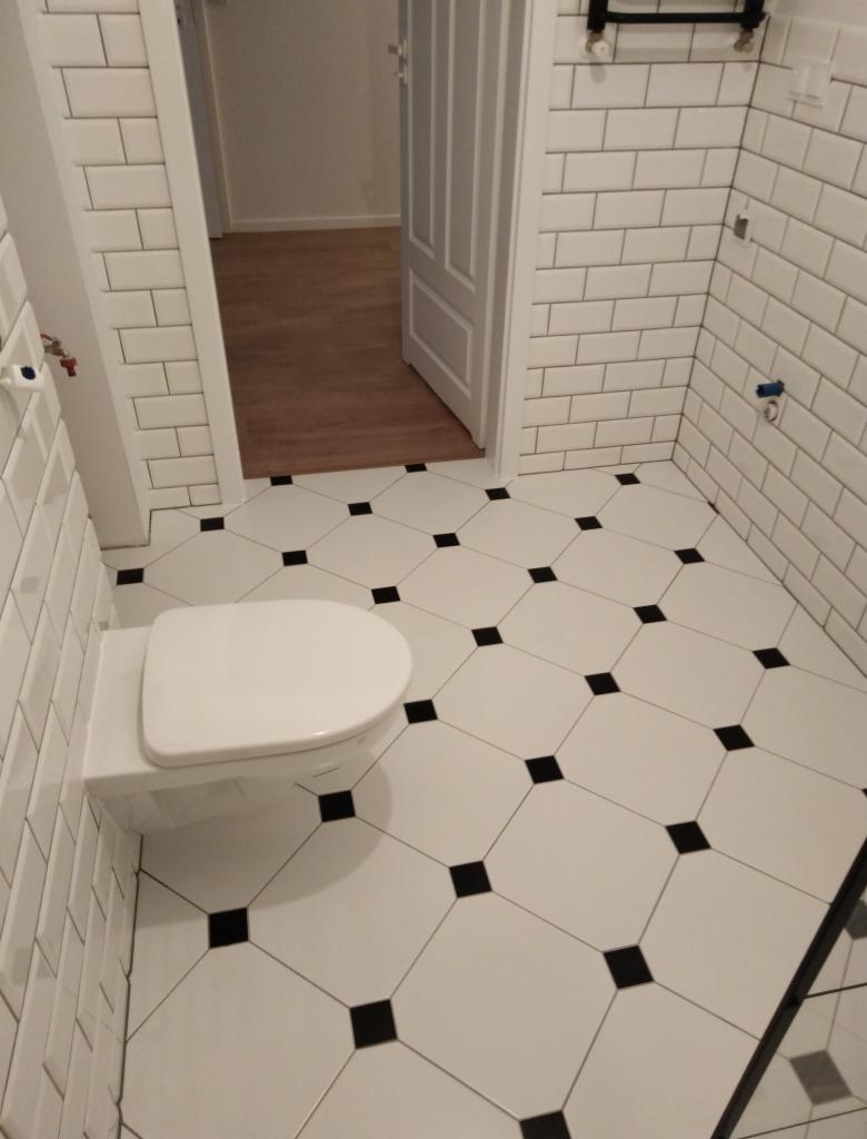 Nowoczesna łazienka z białymi, prostokątnymi płytkami na ścianach i podłodze w biało-czarną szachownicę; wisząca toaleta z deską; widok na otwarte drzwi do innego pomieszczenia z drewnianą podłogą.