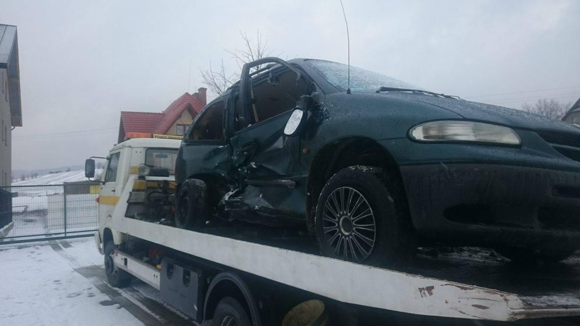 Uszkodzony, zielony minivan załadowany na lawetę w zimowej scenerii, z otwartymi drzwiami i widocznymi wgnieceniami.