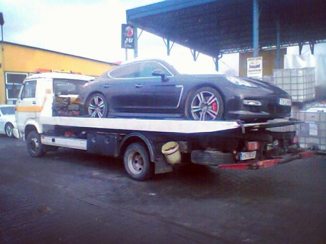 Czarny samochód sportowy marki Porsche na lawecie holowniczej z widocznymi czerwonymi zaciskami hamulcowymi, w tle stacja benzynowa.