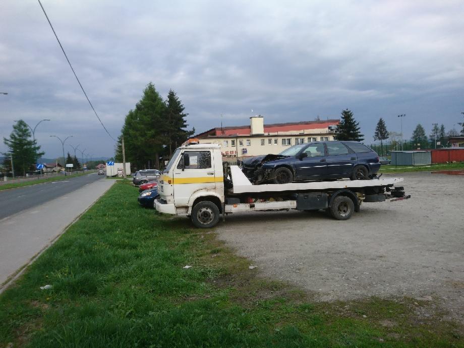 Biała laweta z żółtym pasem transportuje rozbity, ciemny samochód kombi na tle szarego nieba i budynku.