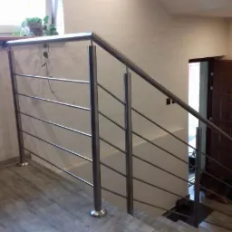 Schody z metalową poręczą i balustradą ze stali nierdzewnej, widok z dołu, w tle doniczka z rośliną na parapecie okna.