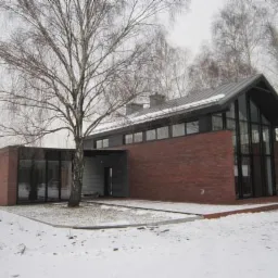 Nowoczesny dom z czerwonej cegły z dużymi oknami i szarym dachem, otoczony śniegiem i drzewami bez liści w zimowej scenerii.