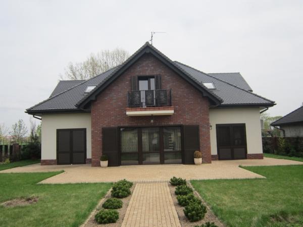 Dwukondygnacyjny dom jednorodzinny z ceglaną fasadą i balkonem, widok z przodu na zadbany trawnik i ścieżkę z kostki.