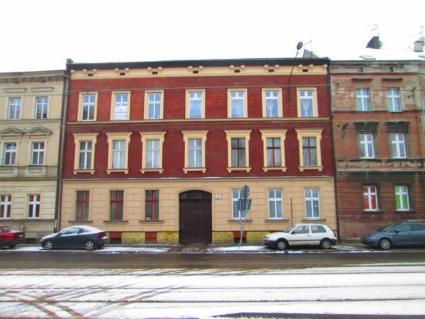 Trzy kamienice w zimowej scenerii, środkowa odnowiona z czerwonej cegły z jasnymi obramowaniami okien, z plakatem w oknie na drugim piętrze.