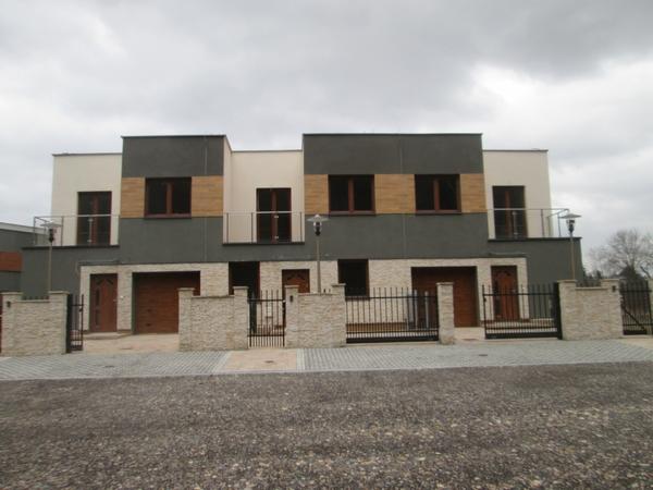 Nowoczesne, dwukondygnacyjne domy w zabudowie szeregowej z garażami, wykończone w odcieniach szarości i bieli, z elementami dekoracyjnymi z kamienia i drewna, ogrodzone kutymi bramami.