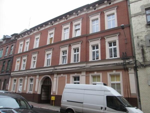 Trzypiętrowy budynek mieszkalny z czerwonej cegły z białymi obramowaniami okien i łukowym przejazdem bramnym, częściowo zasłonięty przez zaparkowany biały samochód dostawczy.