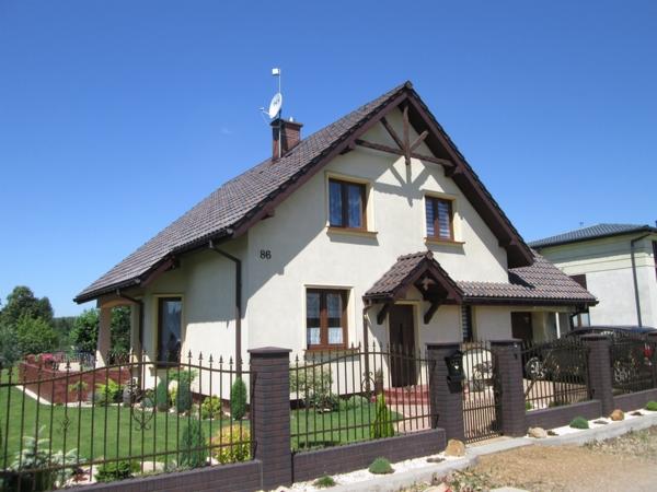 Dom jednorodzinny z beżową elewacją, brązowym dachem i ogrodzeniem z ceglanymi słupkami, numer domu 86 widoczny na ścianie.