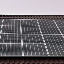 Panel fotowoltaiczny zamontowany na brązowym dachu z blachodachówki, widoczne gołe gałęzie drzew na tle pochmurnego nieba.