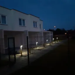 Nowoczesny dom bliźniak oświetlony lampami ogrodowymi wzdłuż ścieżki, widok nocny zza ogrodzenia.