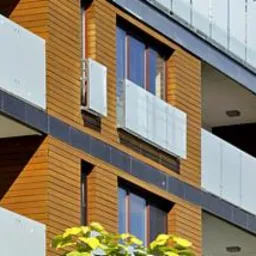 Nowoczesny budynek mieszkalny z balkonami zabezpieczonymi taflami szkła, widok z dołu na fasadę z drewnianą okładziną i ciemnymi elementami wykończeniowymi.