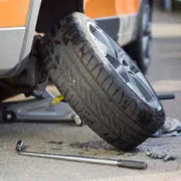 Pomarańczowy samoch&oacute;d na podnośniku z odkręconym kołem, klucz leży na ziemi obok nakrętek.