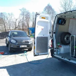 Czyszczenie samochodu Toyota RAV4 z użyciem węża ciśnieniowego; pracownik w kombinezonie roboczym obok pojazdu, z otwartymi drzwiami furgonetki serwisowej w tle.