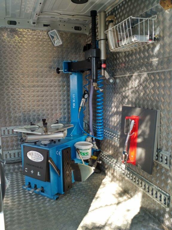 Mobilna montażownica do opon TW X-11 w przestrzeni ładunkowej furgonetki z blachą ryflowaną na ścianach i podłodze, widoczny koszyk z akcesoriami i narzędzia na ścianie.