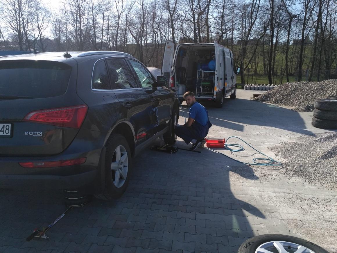 Mechanik w niebieskiej koszulce wymienia koło w ciemnoszarym Audi Q5 na zewnątrz, z otwartym vanem serwisowym w tle i stosem opon obok.