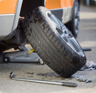 Pomarańczowy samochód na podnośniku z odkręconym kołem, klucz leży na ziemi obok nakrętek.