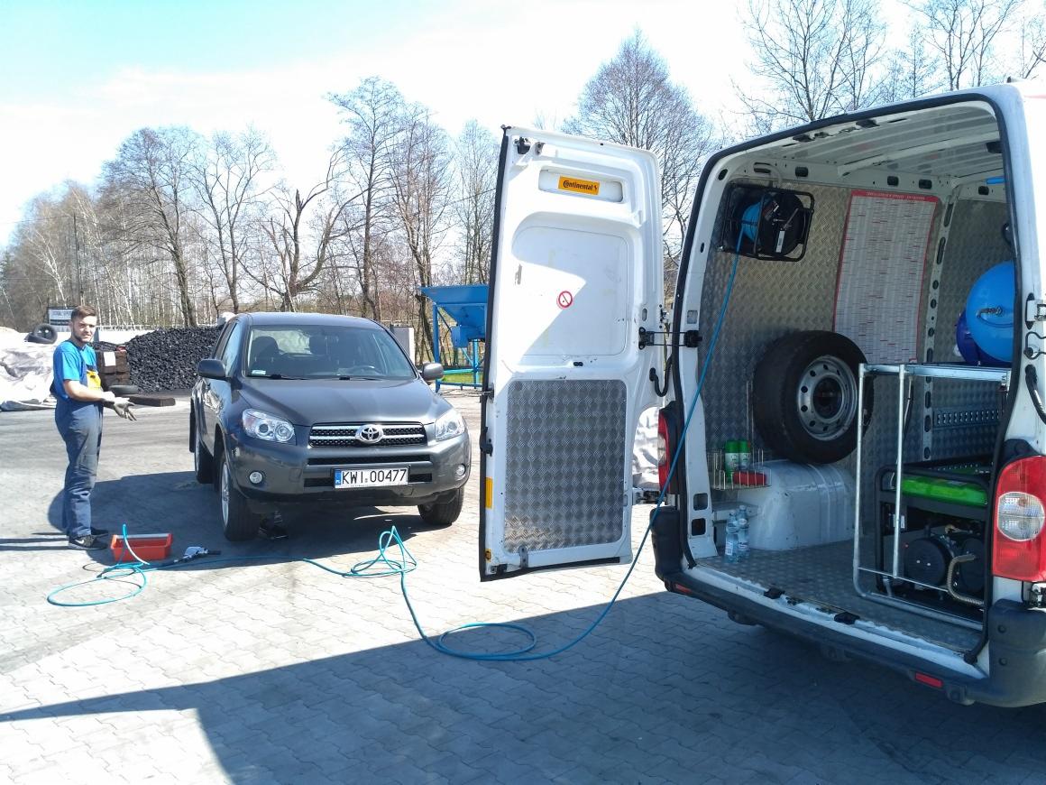 Czyszczenie samochodu Toyota RAV4 z użyciem węża ciśnieniowego; pracownik w kombinezonie roboczym obok pojazdu, z otwartymi drzwiami furgonetki serwisowej w tle.