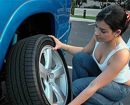 Kobieta w jeansach i białym topie sprawdza oponę z aluminiową felgą przy niebieskim samochodzie na parkingu.