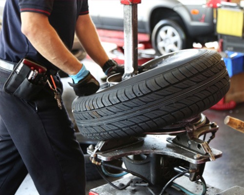Pracownik w niebieskich rękawicach obsługuje maszynę do zmiany opon w warsztacie, w tle widoczny samochód.