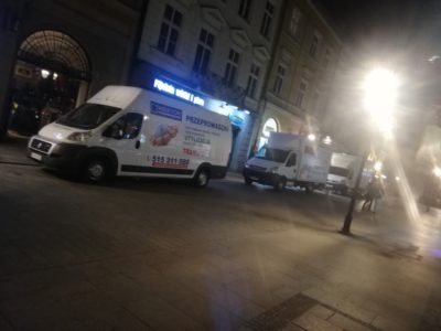 Dwa białe busy firmy transportowej z widocznym numerem telefonu zaparkowane na oświetlonej ulicy miasta nocą.