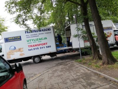 Dwa białe samochody dostawcze firmy przeprowadzkowej Wektor z otwartymi przestrzeniami ładunkowymi podczas załadunku na chodniku w otoczeniu drzew, widoczny numer telefonu i logo firmy.