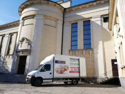 Biały samochód dostawczy z logo firmy przeprowadzkowej zaparkowany przed budynkiem kościoła z wieżyczką