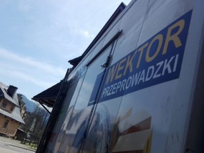 Bok samochodu dostawczego z logo firmy przeprowadzkowej 'WEKTOR Przeprowadzki', widok z dołu na tle błękitnego nieba i fragmentu drewnianego budynku w stylu góralskim.