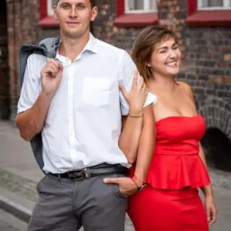 Uśmiechnięta para: mężczyzna w białej koszuli z marynarką przewieszoną przez ramię i kobieta w czerwonej sukience, pozują na tle ceglanej ściany z oknami.