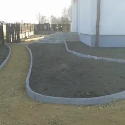 Ukształtowany teren wokół domu z obrzeżami z szarej kostki, ścieżka z jasnego kruszywa prowadząca wzdłuż ogrodzenia i do schodów wejściowych.