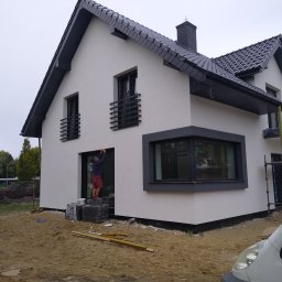 Tomasz Komplet Serwis Firma Ogólnobudowlana - Dwukondygnacyjny dom jednorodzinny z ciemną dachówką i białymi ścianami elewacyjnymi, w trakcie wykańczania; widoczny pracownik na zewnątrz, stojący na bloczkach, poprawiający wykończenie wokół okna.