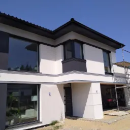 Nowoczesny dom jednorodzinny z białą elewacją i ciemnymi akcentami, widoczne rusztowanie przy garażu, błękitne niebo w tle.