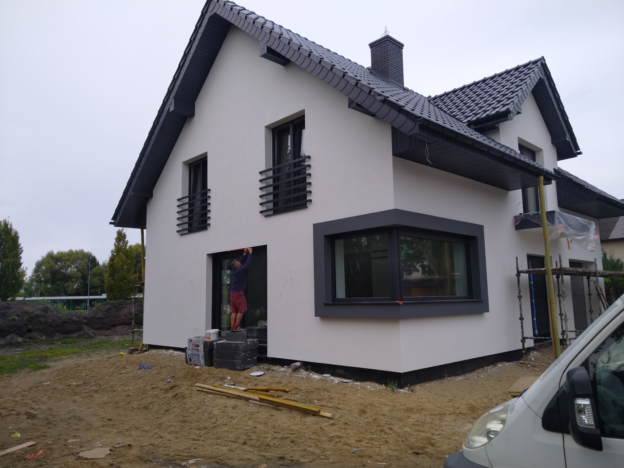 Dwukondygnacyjny dom jednorodzinny z ciemną dachówką i białymi ścianami elewacyjnymi, w trakcie wykańczania; widoczny pracownik na zewnątrz, stojący na bloczkach, poprawiający wykończenie wokół okna.