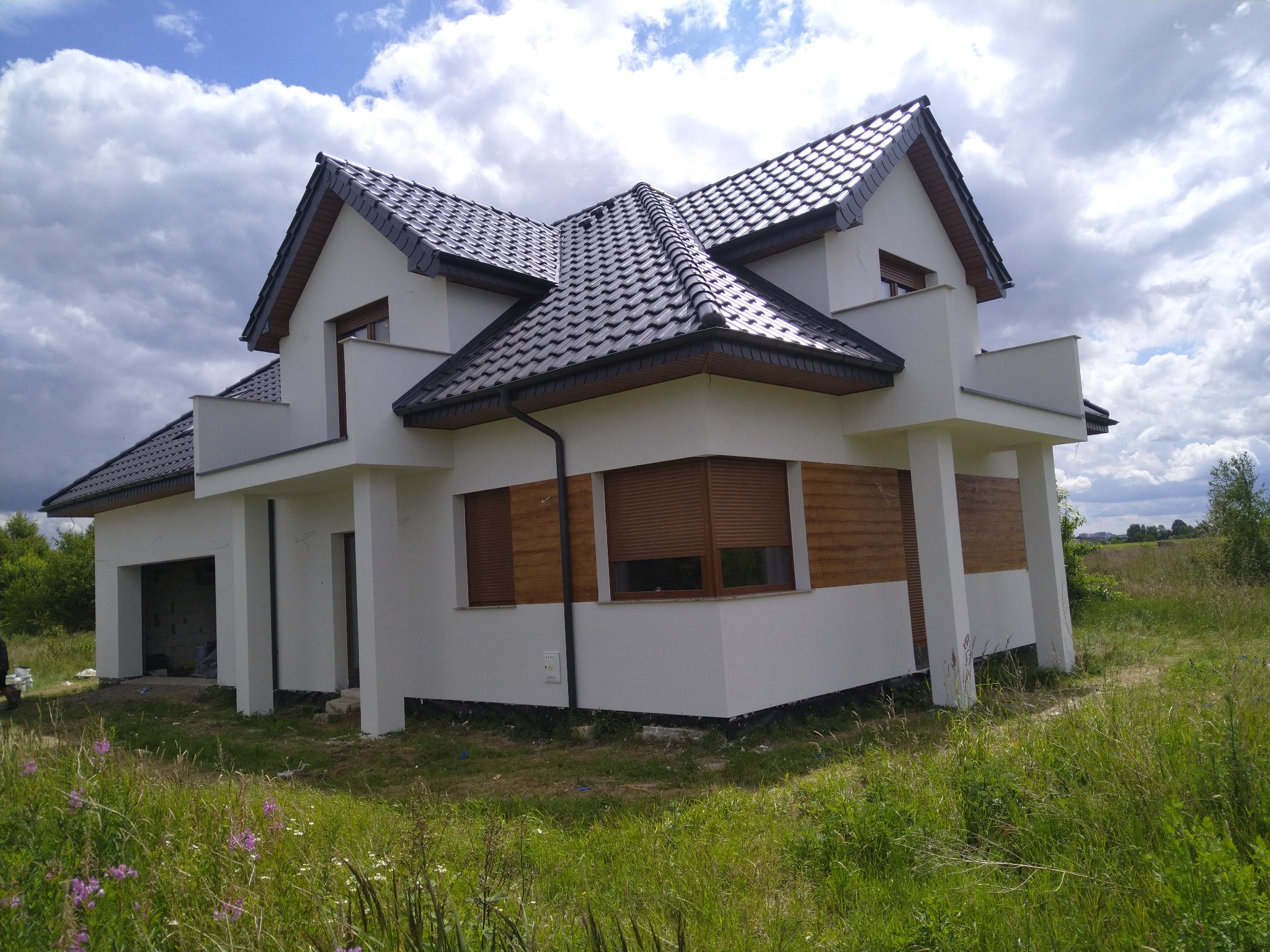 Nowoczesny, dwukondygnacyjny dom z białymi ścianami i ciemną dachówką, otoczony wysoką trawą i kwiatami polnymi, na tle błękitnego nieba z chmurami.