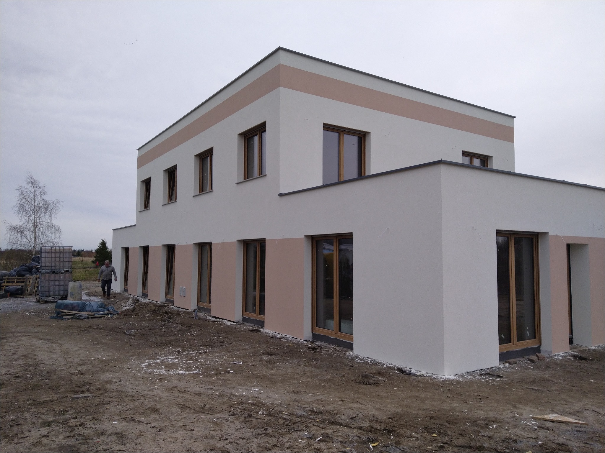 Dwukondygnacyjny budynek z jasną elewacją i poziomym pasem w kolorze beżowym, widoczne okna z drewnianą ramą, teren wokół budynku w trakcie prac wykończeniowych, widoczny mężczyzna i materiały...
