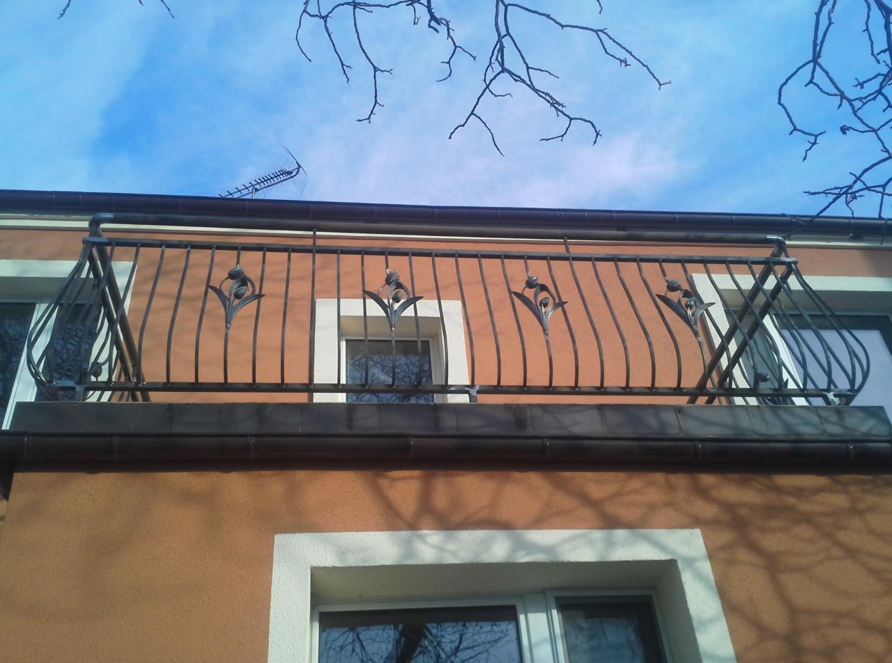 Kuty balkon z dekoracyjnymi elementami w kształcie kwiatów na tle pomarańczowej elewacji i błękitnego nieba z gałęziami drzew