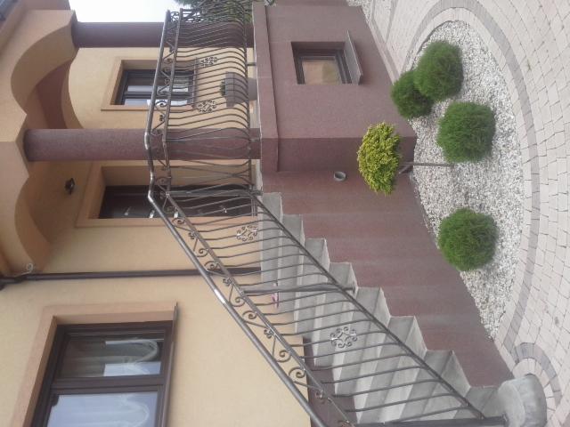 Zewnętrzne schody z metalową balustradą o dekoracyjnych wzorach, prowadzące do budynku z beżową elewacją i brązowymi elementami architektonicznymi, w tle widoczne okna i ozdobne krzewy.
