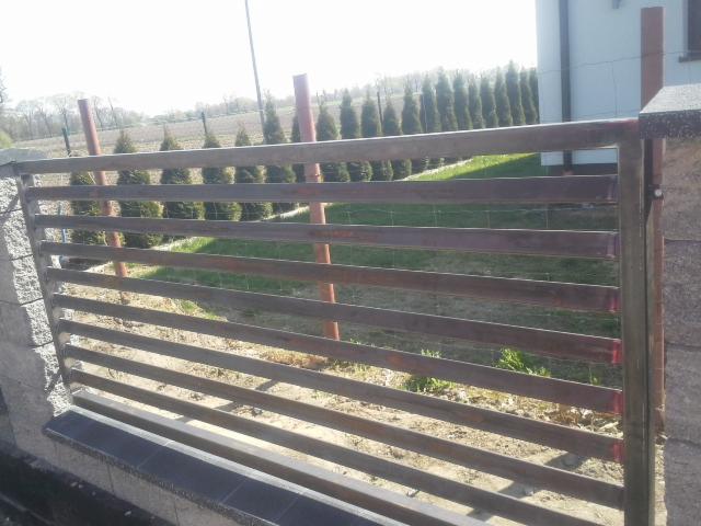 Metalowa brama posesji z poziomymi profilami, widoczne spawy i niedomalowania, w tle rząd tuj i budynek.