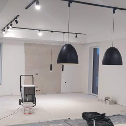 ELPA Instalacje Elektryczne - Montaż oświetlenia szynowego z wiszącymi lampami kloszowymi w surowym wnętrzu, widoczny osuszacz powietrza i narzędzia.