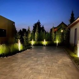 ELPA Instalacje Elektryczne - Oświetlona posesja wieczorową porą, nowoczesne lampy ogrodowe wzdłuż ścieżki i rabat, budynki w tle, spokojna atmosfera.